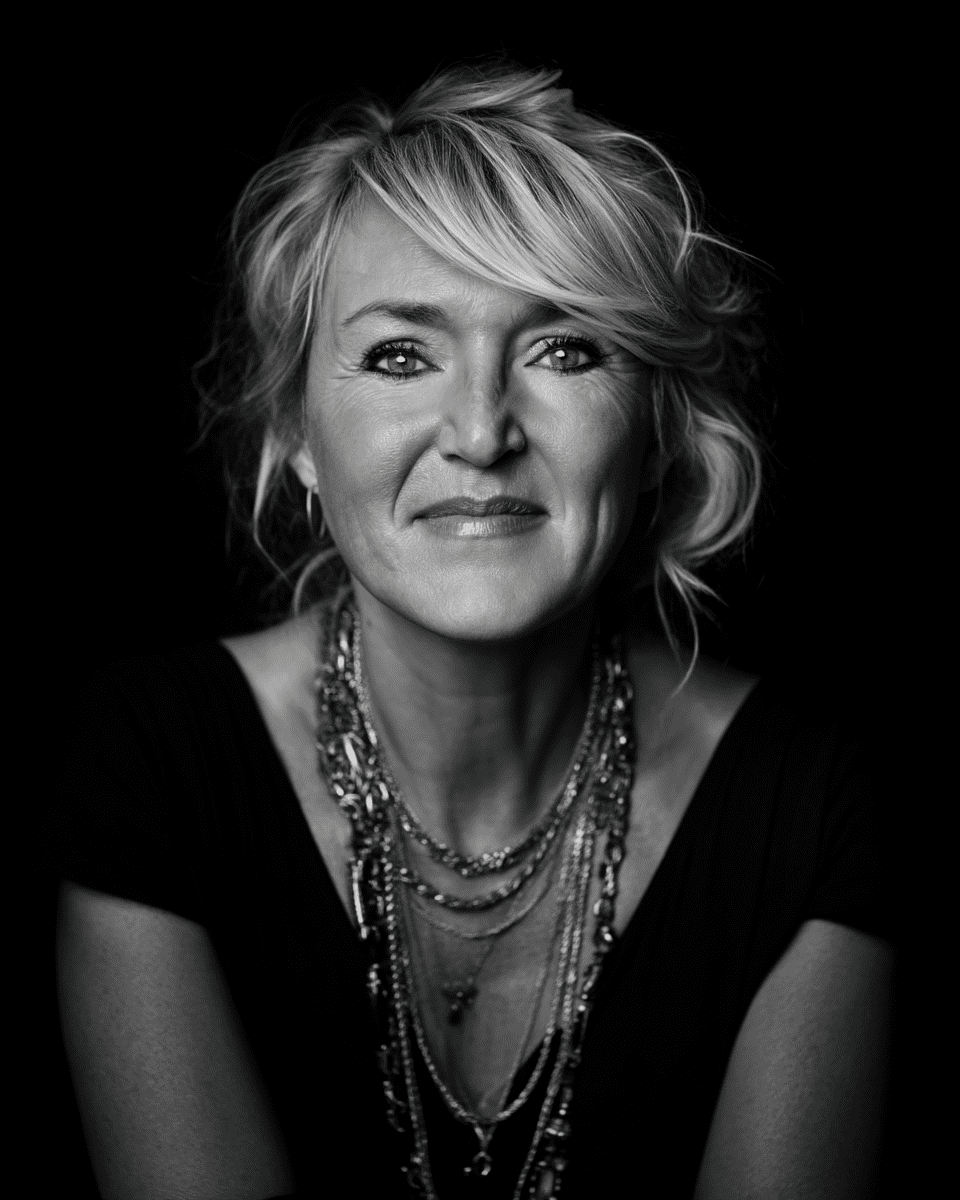 Black and white portrait of a confident mature woman with layered necklaces and soft lighting against a dark background.